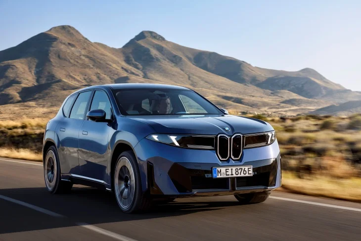Dağlık arazideki asfalt bir yolda dinamik bir şekilde ilerleyen metalik mavi renkli yeni BMW iX3 elektrikli SUV, Neue Klasse tasarım dilinin modern ve minimalist çizgilerini, karakteristik dikey böbrek ızgaraları ve keskin LED farlarıyla yansıtırken markanın elektrikli geleceğini vurguluyor.