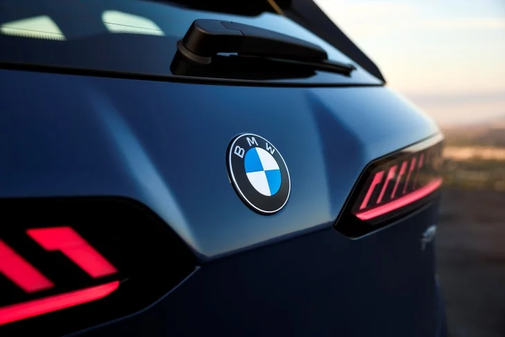 Koyu lacivert renkli yeni BMW iX3'ün sağ arka kısmından çekilmiş bu yakın planda, bagaj kapağındaki parlak BMW logosu, arka cam sileceği ve Neue Klasse tasarım dilinin bir parçası olan modern çizgilerle şekillendirilmiş karakteristik kırmızı LED stop lambası vurgulanıyor, elektrikli SUV'un güçlü ve teknolojik geleceğini yansıtıyor.