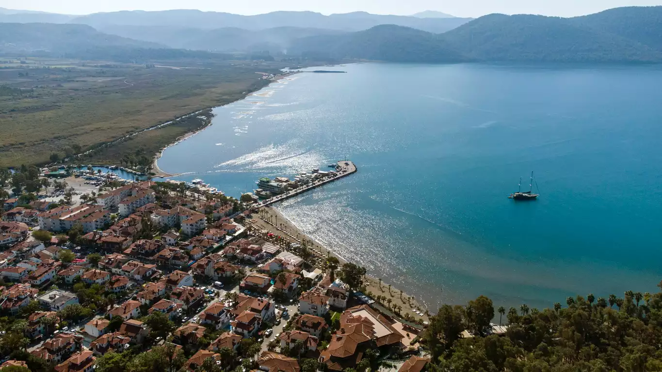 Akyaka'nın kuşbakışı görüntüsü, kırmızı çatılı evlerin sahil boyunca uzandığı, yatların demirlediği bir marina ve uzun bir iskelenin berrak turkuaz sulara açıldığı, arka planda yeşil dağların yükseldiği güneşli bir koyu gösteriyor.