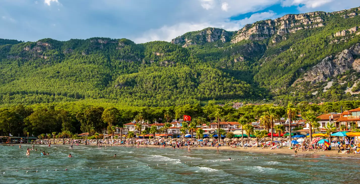 Akyaka'nın berrak sularında yüzen ve sahilde güneşlenen tatilcilerle dolu canlı plajı, yemyeşil ormanlarla kaplı heybetli dağların eteklerinde, geleneksel evler ve palmiye ağaçlarıyla çevrili bir yaz gününü gözler önüne seriyor.