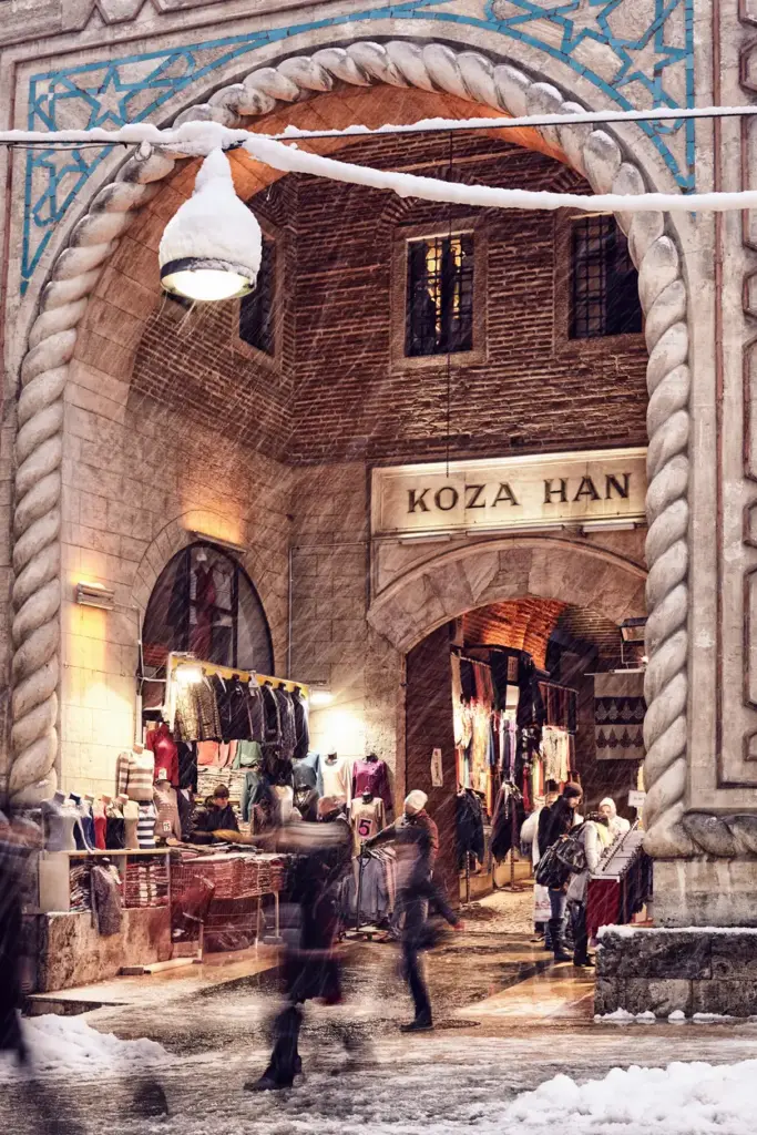 Bursa'nın önemli gezilecek yerlerinden Koza Han'ın karla kaplı görkemli taş kemerli girişinde, hafifçe yağan karlar altında hareketli bir ticaret ve sosyal yaşam süren insan silüetleri, hanın asırlık ruhunu ve şehrin Osmanlı mirasını yansıtıyor.
