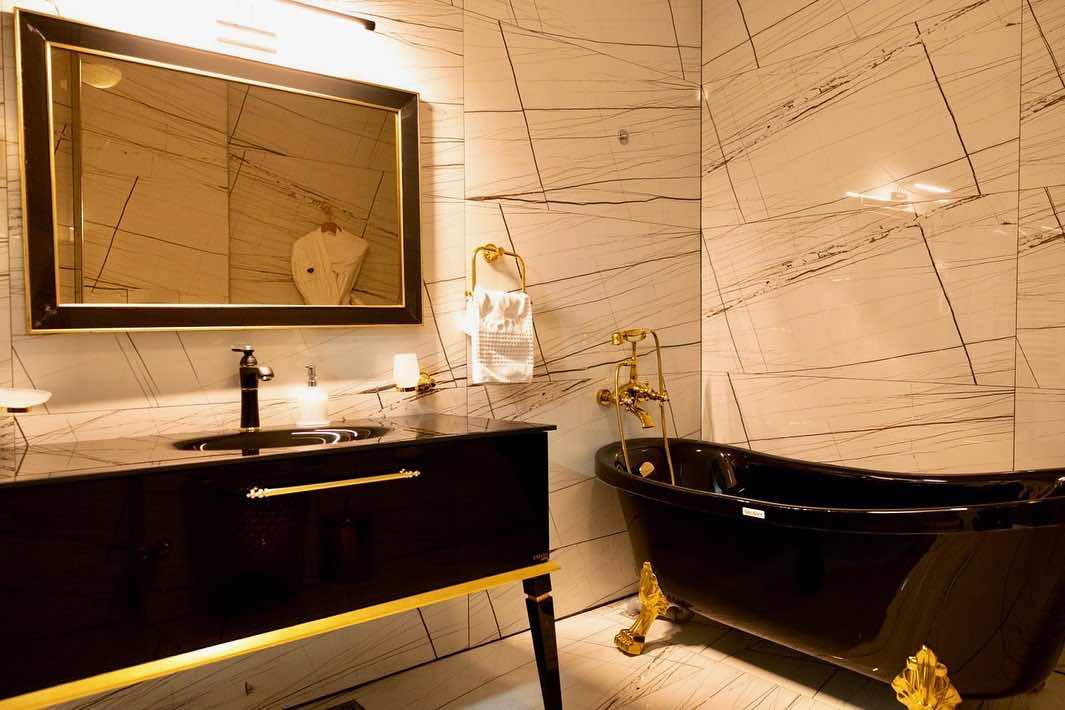 Bir Üsküp seyahati sırasında konforlu bir konaklama deneyimi sunan Bushi Resort & Spa'nın, siyah lavabo dolabı ve altın detaylarla modern lüksü yansıtan, altın ayaklı siyah küveti ve damarlı mermer duvarlarıyla şık bir banyosu.