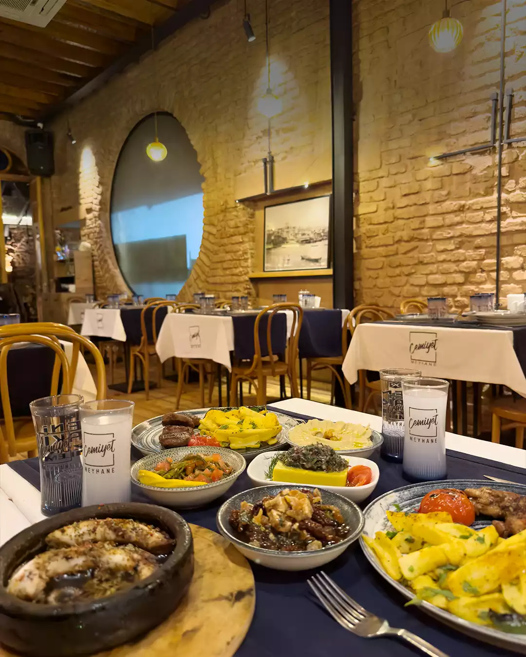 Cemiyet Meyhanesi'nde, tuğla duvarlı, sıcak atmosferli bir ortamda, zengin meze çeşitleri, çömlekte balık ve et yemekleriyle donatılmış lacivert örtülü bir masada iki kadeh rakı, İstanbul'un en iyi meyhane deneyimlerinden birini sunuyor.