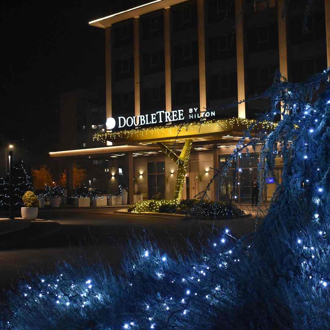Üsküp seyahat rehberi kapsamında konaklama seçeneklerinden biri olan DoubleTree by Hilton Skopje otelinin gece çekilmiş, girişindeki parlak sarı ve beyaz ışıklarla aydınlanmış tabelası ve süslü çalılarla davetkar görünen ön cephesi.