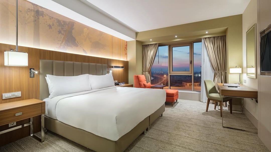 Üsküp'teki konforlu bir konaklama deneyimi sunan modern ve ferah bir otel odasında, beyaz çarşaflı geniş yatak, pencere kenarında turuncu tekli koltuk ve puf, çalışma masası ile şehir ışıklarının göründüğü akşam manzarası eşliğinde şık bir atmosfer.