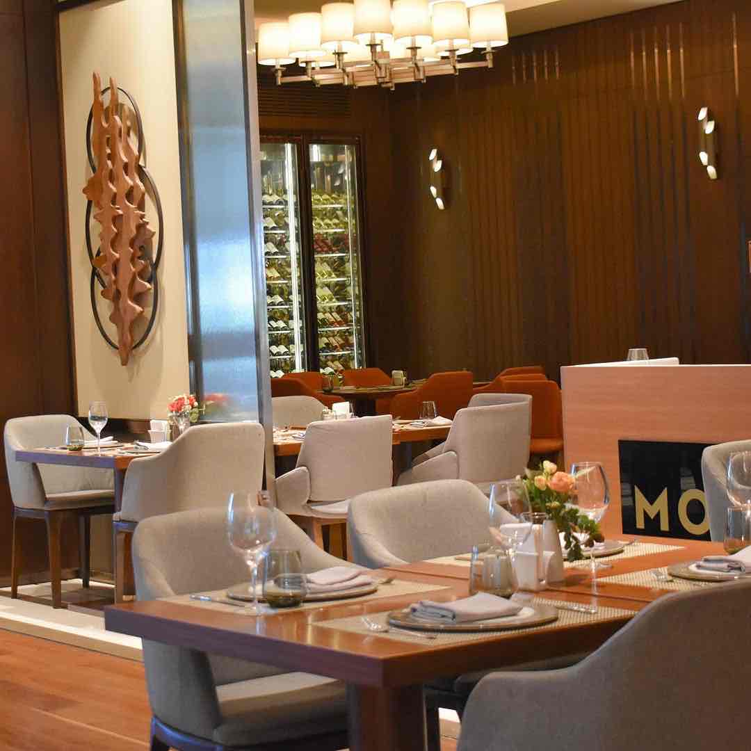 DoubleTree by Hilton Skopje'nin şık Monte Restoranı'nda, özenle düzenlenmiş ahşap masalar, krem renkli sandalyeler, modern avize, duvarda soyut ahşap sanat eseri ve arka planda bir şarap dolabıyla Üsküp seyahatinde konforlu bir gastronomi deneyimi sunan çağdaş ve lüks bir yemek alanı.