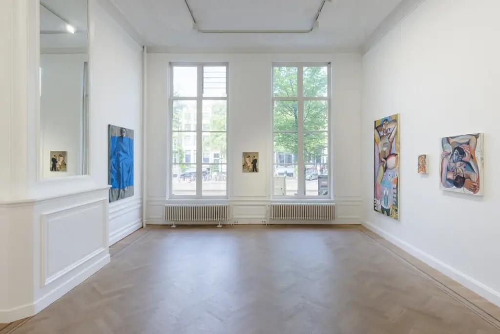 Amsterdam'ın çağdaş sanat sahnesine atıfta bulunan bu geniş ve aydınlık galeride, beyaz duvarlarda asılı soyut ve figüratif tablolar, herringbone parke zemini ve dışarıdaki yeşil ağaçları gösteren büyük pencereler modern bir sergi atmosferi sunuyor.