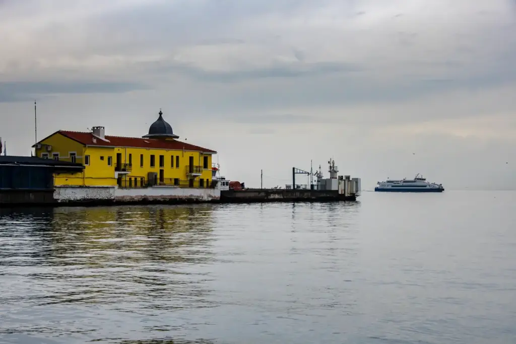 Bulutlu gri bir gökyüzünün altında, sakin denizde sarı renkli, kırmızı çatılı ve kubbemsi bir çatıya sahip eski bir iskele binası, hemen yanındaki gri rıhtım ve uzakta seyreden beyaz bir feribotun panoramik görüntüsü.