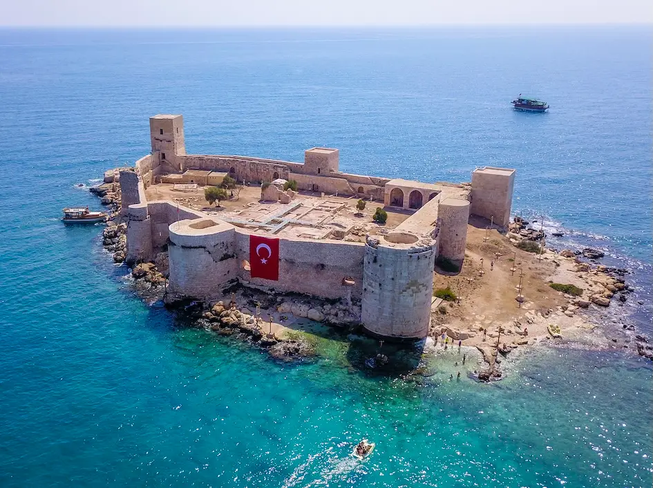 Mersin'de gezilecek yerler arasında öne çıkan, masmavi Akdeniz'in ortasında küçük bir adacık üzerine kurulmuş, surları ve kuleleriyle tarihi bir yapı olan Kızkalesi'nin havadan görünüşü; kale duvarında asılı büyük Türk bayrağı, çevresindeki turkuaz sularda yüzen insanlar ve kıyıya yanaşmış teknelerle Mersin kıyı serüvenini yansıtıyor.