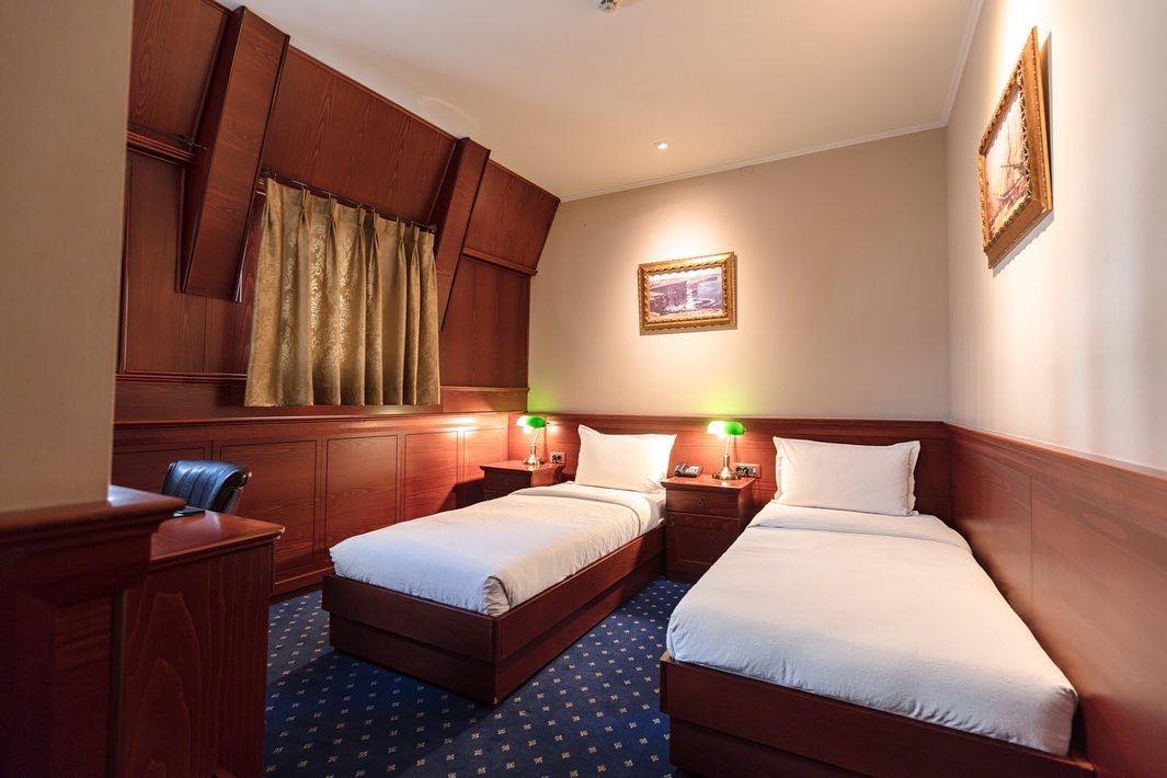 Üsküp seyahat rehberi kapsamında incelenen Hotel Senigallia'nın karakterli odalarından birinde, koyu ahşap panellerle çevrili alanda konumlanmış iki tek kişilik yatak, bej duvarlar, yeşil başlıklı abajurlar ve zarif tablolar dikkat çekiyor.