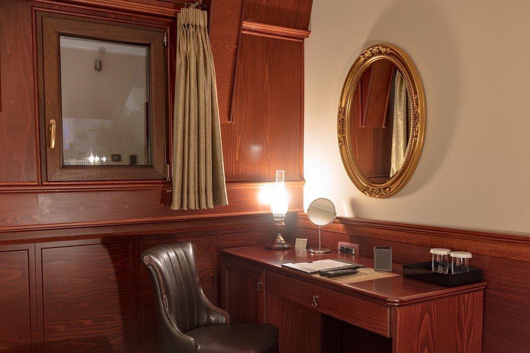 Koyu renk ahşap panellerle döşenmiş, klasik tarzda dekore edilmiş bir Üsküp otel odasında; masa lambası, küçük makyaj aynası, iki kadeh bulunan tepsi ve evrakların yer aldığı ahşap bir çalışma masası, yanında kahverengi deri sandalye ve duvarda altın çerçeveli oval bir ayna ile odanın zarif konaklama atmosferi.