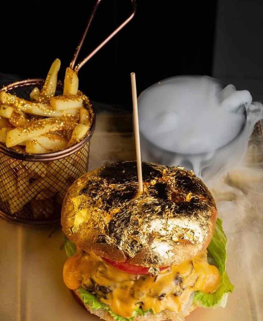 Üsküp'ün modern gastronomi sahnesinin dikkat çekici bir örneği olarak sunulan, üzeri altın varakla kaplı gösterişli bir hamburger ve yanında simli patates kızartmaları, sisli bir arka plan eşliğinde şık bir sunumla iştah açıcı görünüyor.