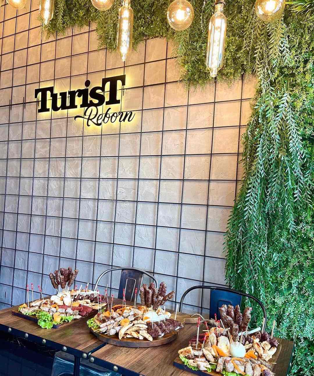 Üsküp'teki Turist Reborn restoranının modern iç mekanında, ızgara etler, köfteler ve çeşitli mezelerle dolu ahşap masalar, tavandan sarkan sıcak ampuller ve sağda yemyeşil bitki duvarıyla, şehrin seyahat rehberi deneyimine otantik Makedon lezzetlerini taşıyan davetkar bir atmosfer sunuluyor.