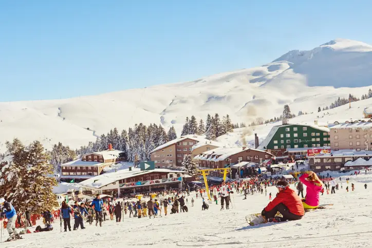 Güneşli bir kış gününde, karla kaplı Uludağ'ın zirvesinde, çok sayıda kayakçı ve snowboardcunun pistlerde keyifli anlar yaşadığı, çevredeki modern otellerin ve çam ağaçlarının bembeyaz örtüyle kaplandığı bu canlı manzara, Bursa'da gezilecek yerlerden biri olan popüler kış sporları merkezinin dinamik atmosferini yansıtıyor.