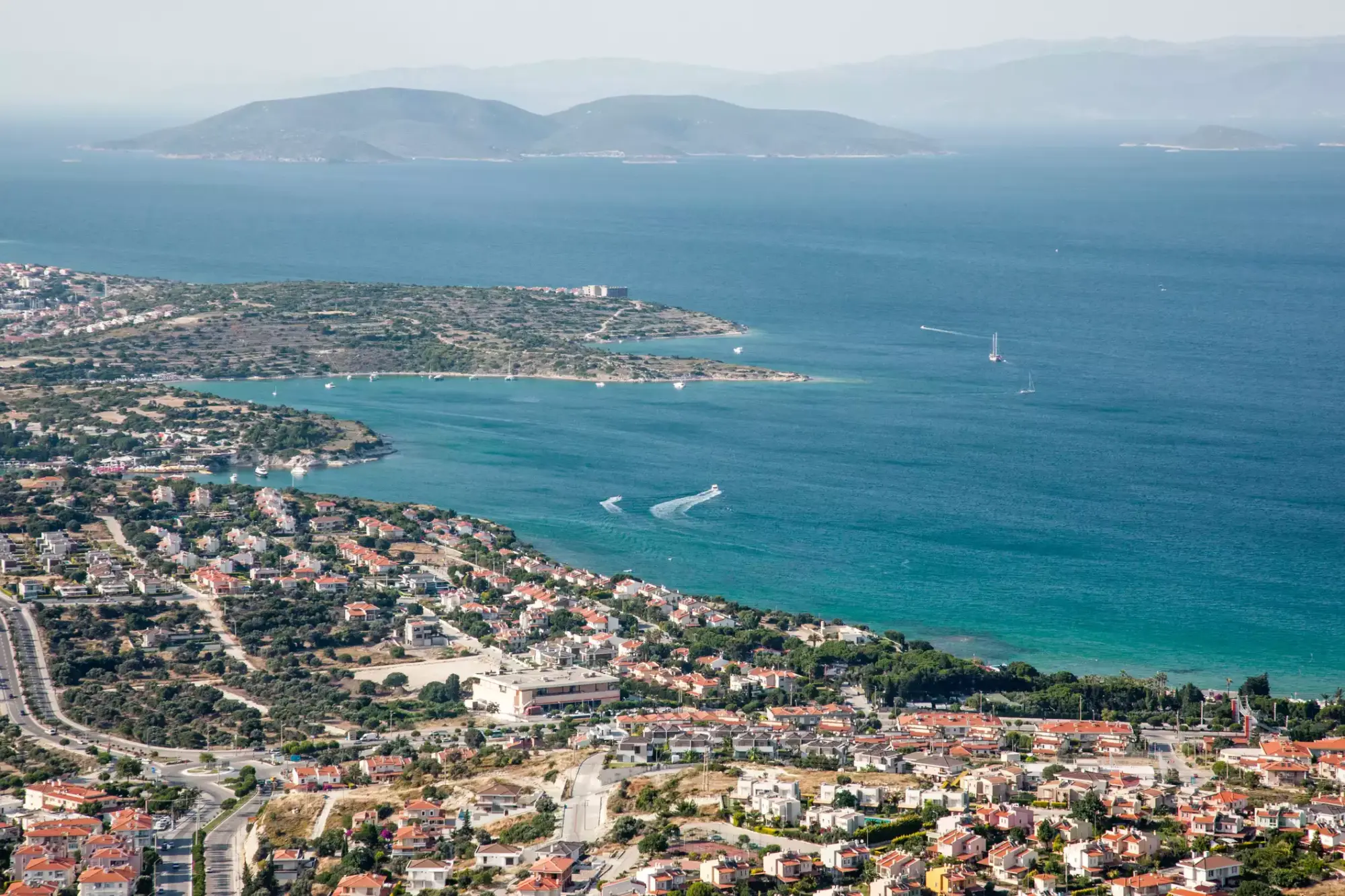Geniş açılı bir hava fotoğrafında, Urla'nın kıvrımlı sahil şeridi boyunca uzanan, kırmızı çatılı evlerin ve yemyeşil ağaçların harmanlandığı yerleşim yerleri, pırıl pırıl turkuaz renkteki Ege Denizi ile iç içe geçiyor; uzakta sisli dağlar ve denizde seyreden tekneler bu huzurlu manzarayı tamamlıyor.