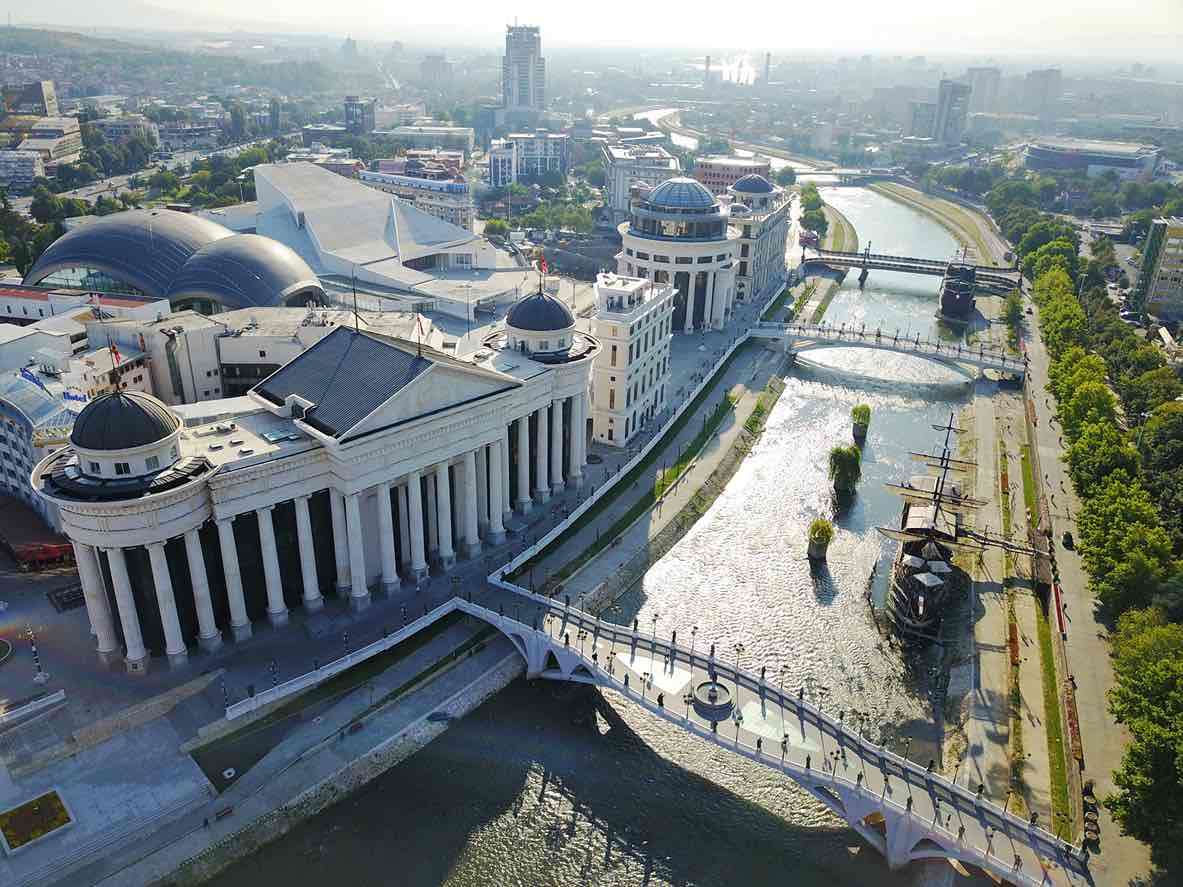 Üsküp'ün merkezinden geçen Vardar Nehri'nin kuşbakışı görünümünde, modernizasyon projeleriyle inşa edilmiş, sütunlu ve kubbeli gösterişli yapılar, heykellerle bezeli köprüler ve nehirdeki gemi replikaları, şehrin zengin ve katmanlı tarihinden kalan izleriyle bugünkü karmaşık yapısını gözler önüne seriyor.