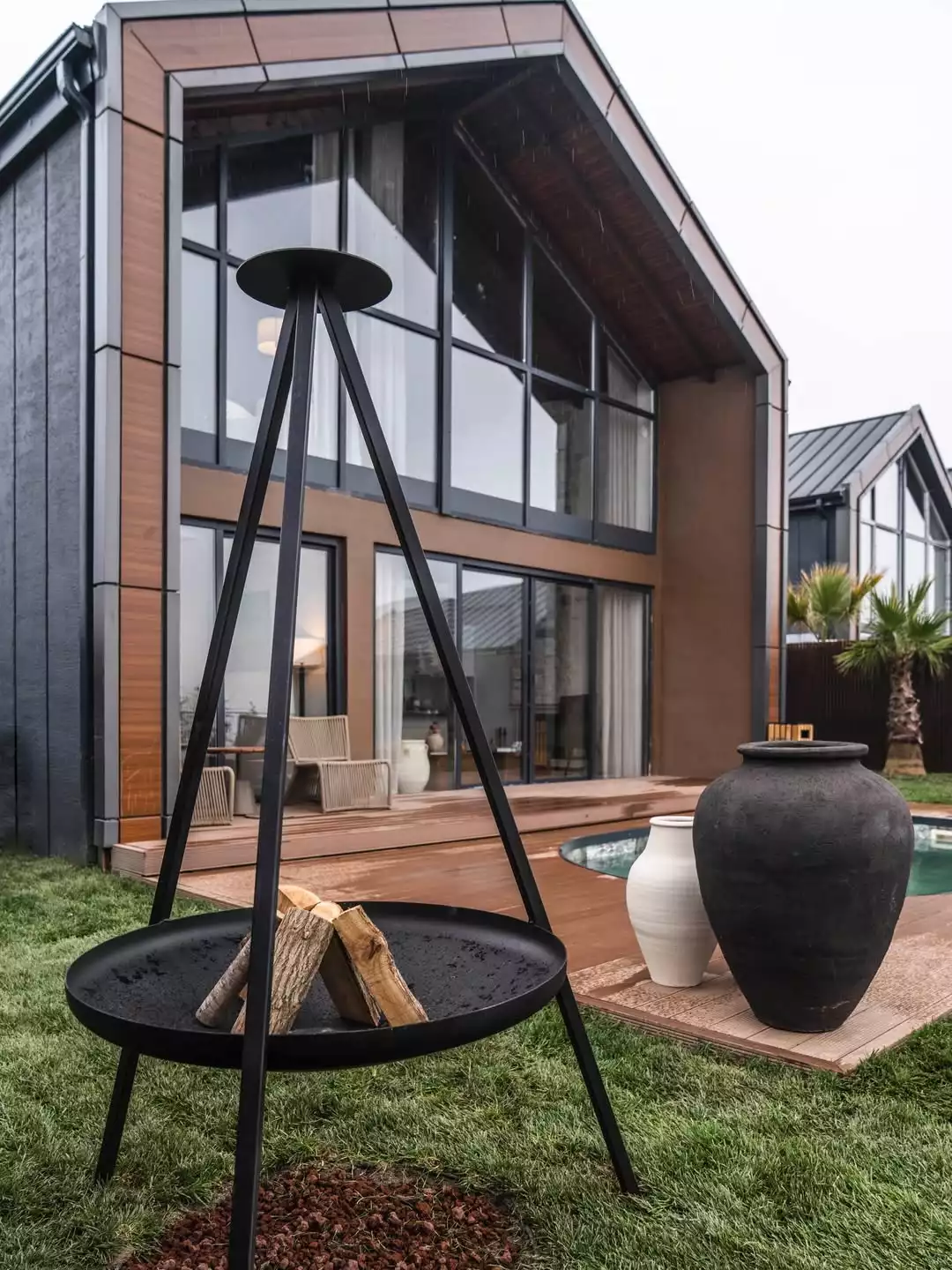 Geniş cam pencereler ve koyu renk ahşap panellerle modern bir mimariye sahip, çarpıcı bir konut yapısının önünde, yeşil çimlerin üzerinde duran siyah, üç ayaklı bir ateş çukuru içinde odunlar bulunurken, arka planda ahşap bir teras, dış mekan oturma grubu, bir havuz ve dekoratif vazolarla tamamlanan davetkar bir yaşam alanı görülüyor.