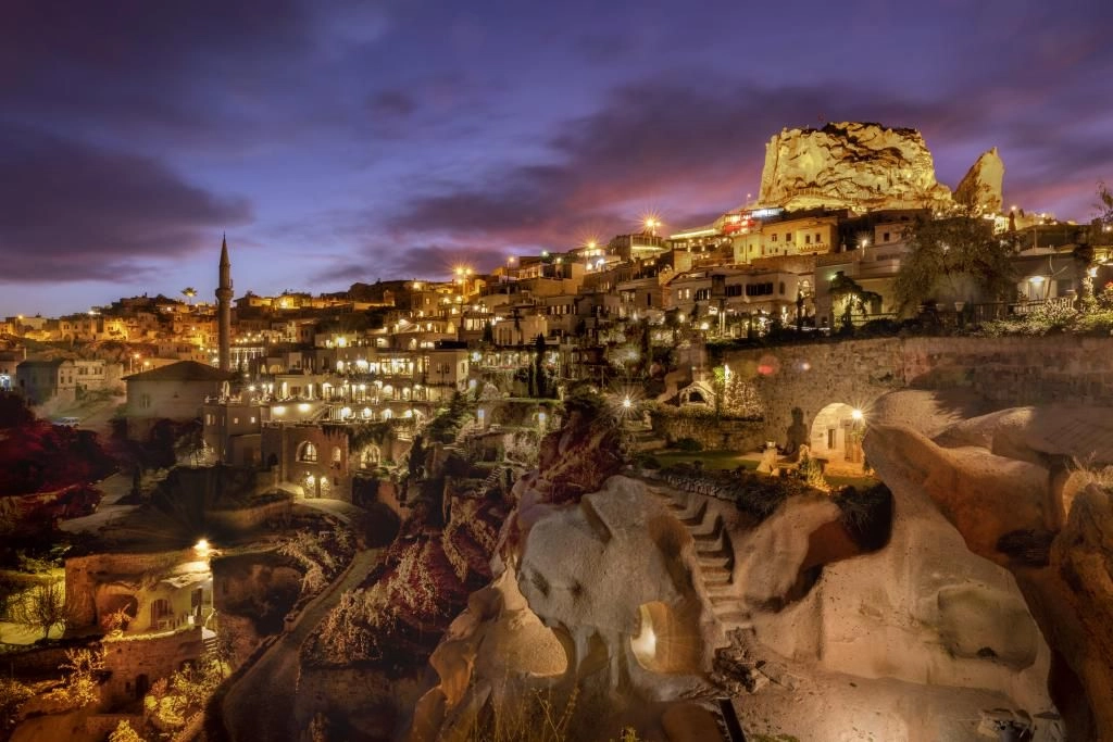 Kapadokya'da bulunan ve 2026 Michelin Anahtarları ile ödüllendirilen Argos in Cappadocia'dan bir otel odası görünümü; doğal taş dokulu duvarları, lüks beyaz nevresimli geniş yatağı ve otantik detaylarıyla dikkat çeken bu özel tasarım oda, misafirlerine benzersiz bir konaklama deneyimi sunuyor.