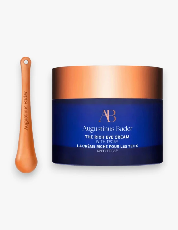 Beyaz arka planda, metalik bakır renkli kapaklı lacivert Augustinus Bader The Rich Eye Cream kavanozu ve yanında duran aynı tonlarda bir uygulama aparatı, göz çevresi bakımı için bir ürün setini detaylıca gösteriyor.