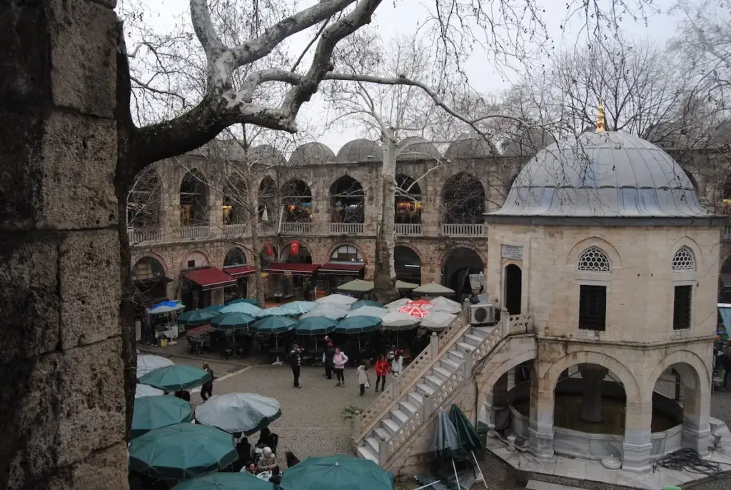 Bursa'nın tarihi Hanlar Bölgesi'ndeki Koza Han'ın, dalları çıplak asırlık çınar ağaçlarıyla çevrili avlusunda, kubbeli şadırvanın etrafındaki iki katlı Osmanlı revakları altında, şemsiyelerin gölgesinde oturan insanlar şehrin canlı sosyal ve ticari ruhunu yansıtır.