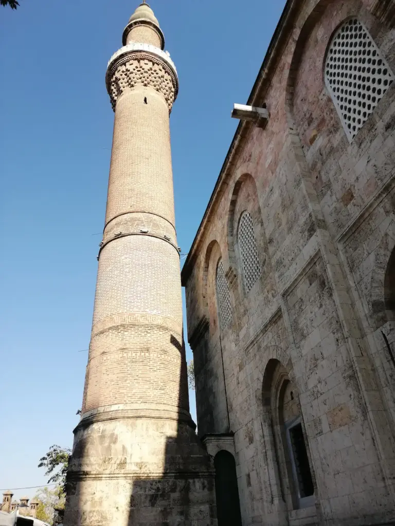 Güneşli bir mavi gökyüzü altında, Osmanlı'nın ilk başkenti Bursa'da gezilecek yerler arasında öne çıkan tarihi Ulu Cami'nin tuğla minaresi, şerefesi altındaki detaylı işçiliği ve sağ yanda yükselen kemerli pencerelere sahip taş duvarlı anıtsal mimarisiyle şehrin kadim ruhunu yansıtıyor.