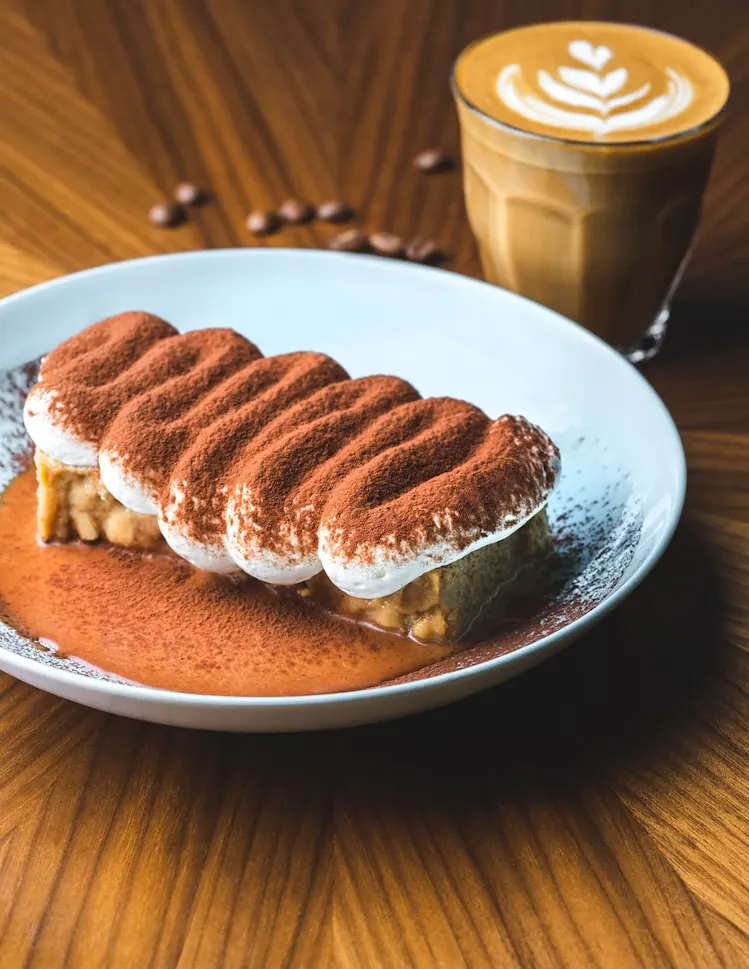 Beyaz bir tabakta, bol kakao tozu serpilmiş, dalgalı kremalı üst katmanıyla öne çıkan, altında karamel soslu bir tatlı; arka planda zarif yaprak desenli latte art'a sahip bir cappuccino ve ahşap masaya özenle dağılmış birkaç kahve çekirdeğiyle tamamlanan, keyifli bir sunum.