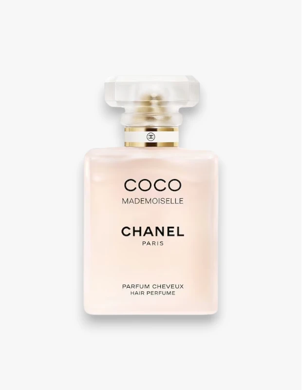Beyaz bir arka plan önünde dikey olarak konumlandırılmış, şeffaf, çok yüzeyli bir kapak ve altın rengi Chanel logosu içeren bir bantla tamamlanmış, üzerinde Coco Mademoiselle ve Parfum Cheveux Hair Perfume yazıları bulunan, soluk pembe renkli şık bir Chanel Coco Mademoiselle saç parfümü şişesi.