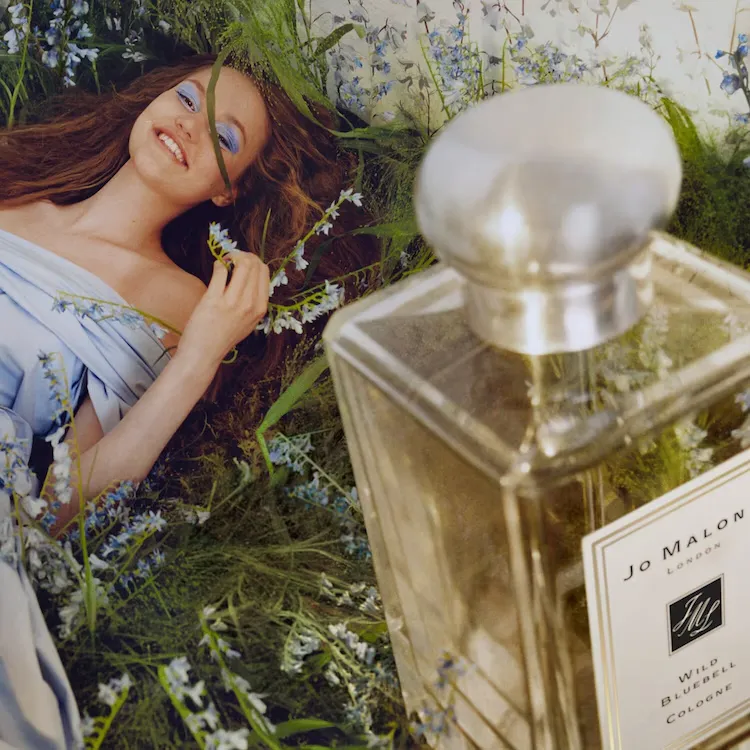 Ön planda hafifçe bulanık Jo Malone London Wild Bluebell Cologne parfüm şişesi, arka planda mavi göz makyajlı ve açık renk elbisesiyle yabani mavi çan çiçekleri ve yemyeşil otların arasına uzanmış gülümseyen bir kadının yer aldığı, doğanın ferahlığını yansıtan bir görsel.