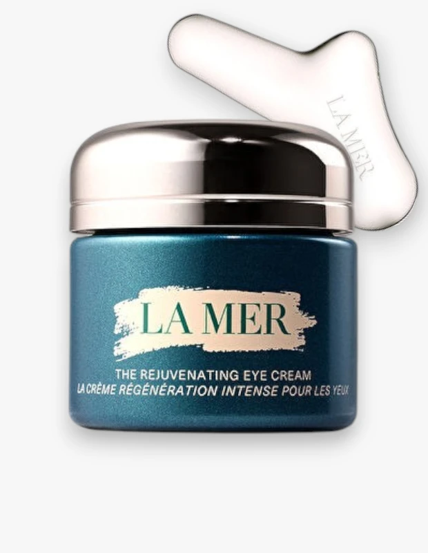 Gümüş renkli parlak kapağı olan, koyu turkuaz bir kavanoz içinde La Mer The Rejuvenating Eye Cream gençleştirici göz kremi ve ürünle birlikte gelen La Mer markalı beyaz bir masaj aleti beyaz bir zeminde dikkat çekici bir şekilde konumlandırılmıştır.