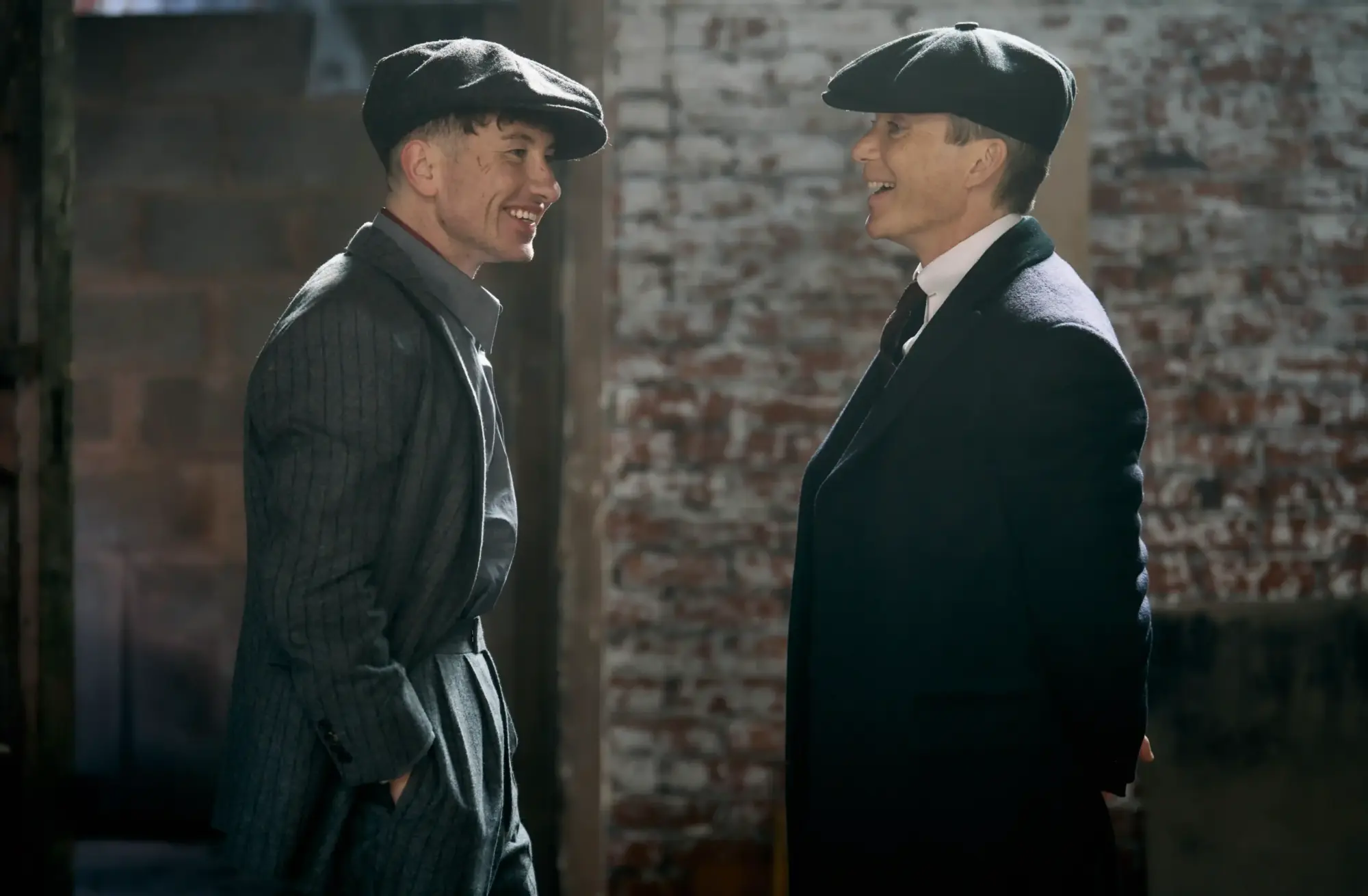 The Immortal Man filminin setinde, 1920'lerin gentleman estetiğini yansıtan kasketleri ve klasik giyim tarzlarıyla Peaky Blinders dünyasından Barry Keoghan ile Cillian Murphy'nin gülümseyerek karşılıklı durduğu, dönem ruhunu taşıyan tuğla duvarlı bir ortamda çekilmiş atmosferik bir kare.