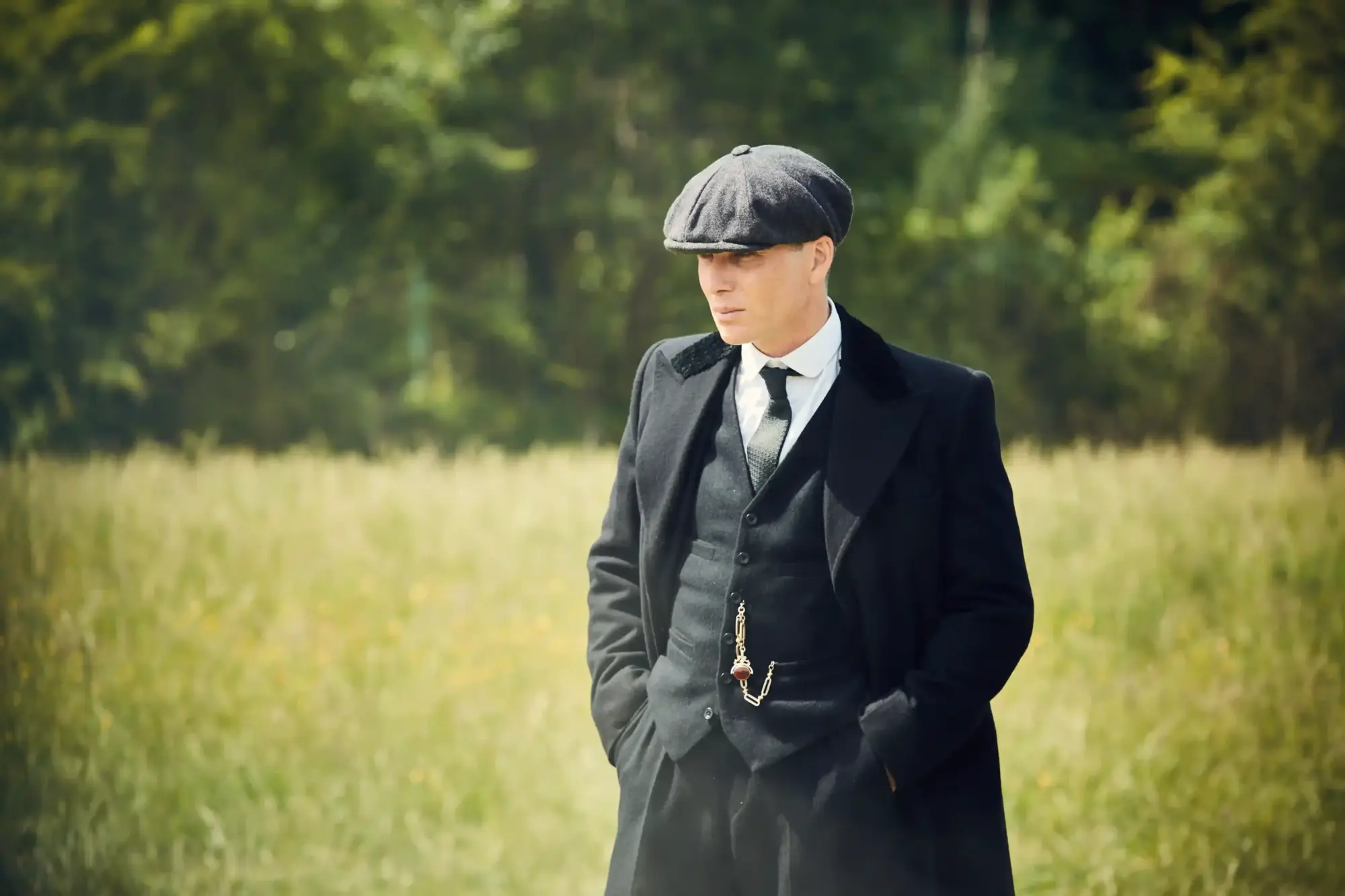 Peaky Blinders’ın ikonik karakteri Thomas Shelby, gri yelekli üç parçalı takım elbisesi, siyah paltosu, kasketi ve cep saati zinciriyle 1920’lerin maskülen zarafetini modern erkek modasına yansıtan keskin bakışıyla yeşil bir dış mekanda duruyor.