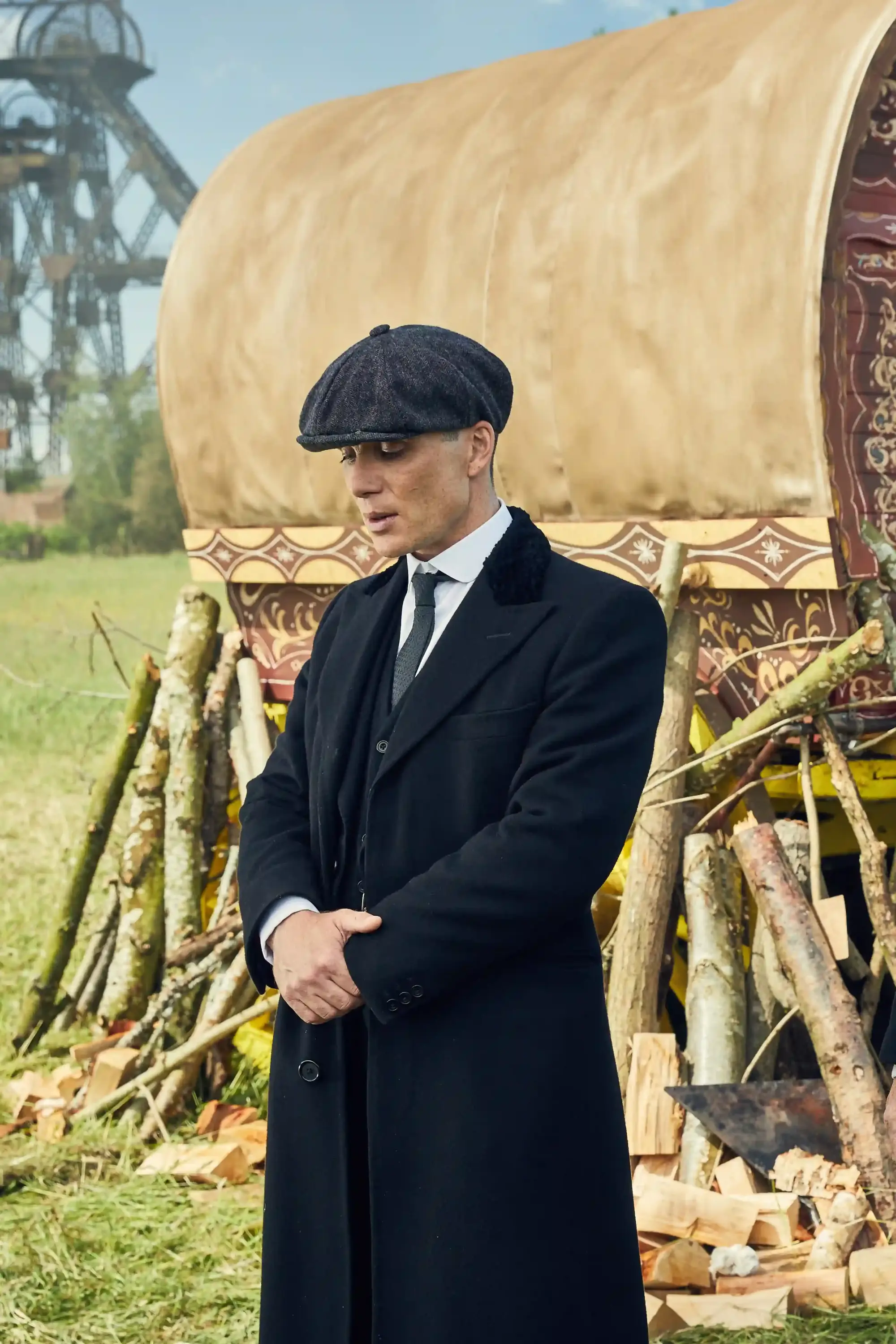 Peaky Blinders dizisindeki Thomas Shelby karakterini canlandıran Cillian Murphy, koyu renk paltosu, gri kasketi ve şık takım elbisesiyle, 1920’lerin maskülen zarafetini ve erkek modasını yansıtan bir karavanın önünde ellerini kavuşturmuş, aşağı bakıyor.