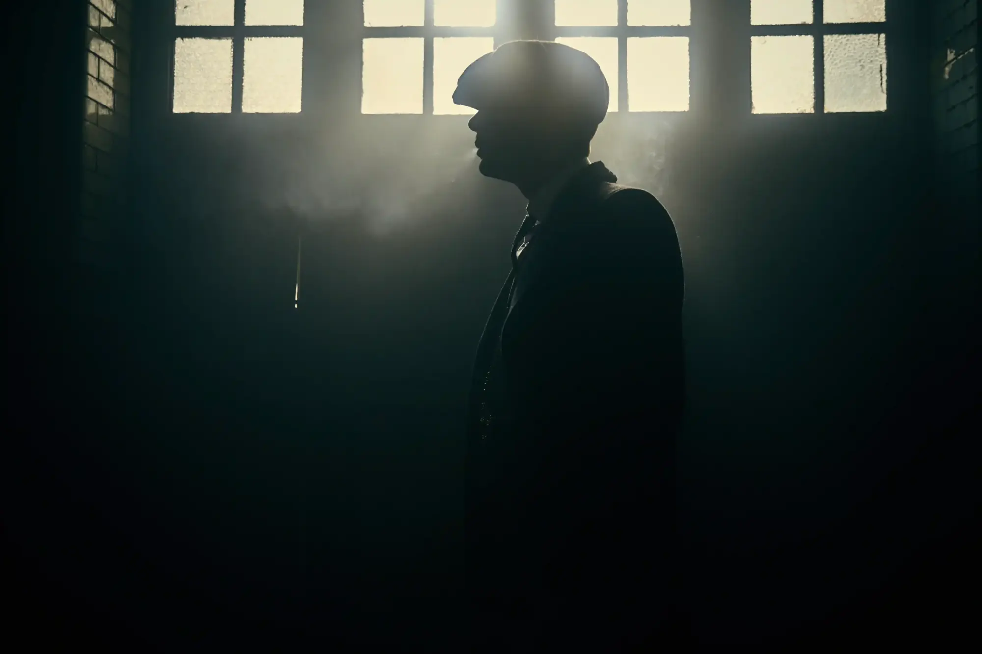 Pencerelerden sızan dramatik ışığın önünde, Peaky Blinders'ın ikonik karakteri Thomas Shelby, kasketi ve zarif takım elbisesiyle gücünü ve gizemini yansıtan etkileyici bir silüet oluşturarak 1920'lerin maskülen zarafetini yeniden gündeme getiriyor.