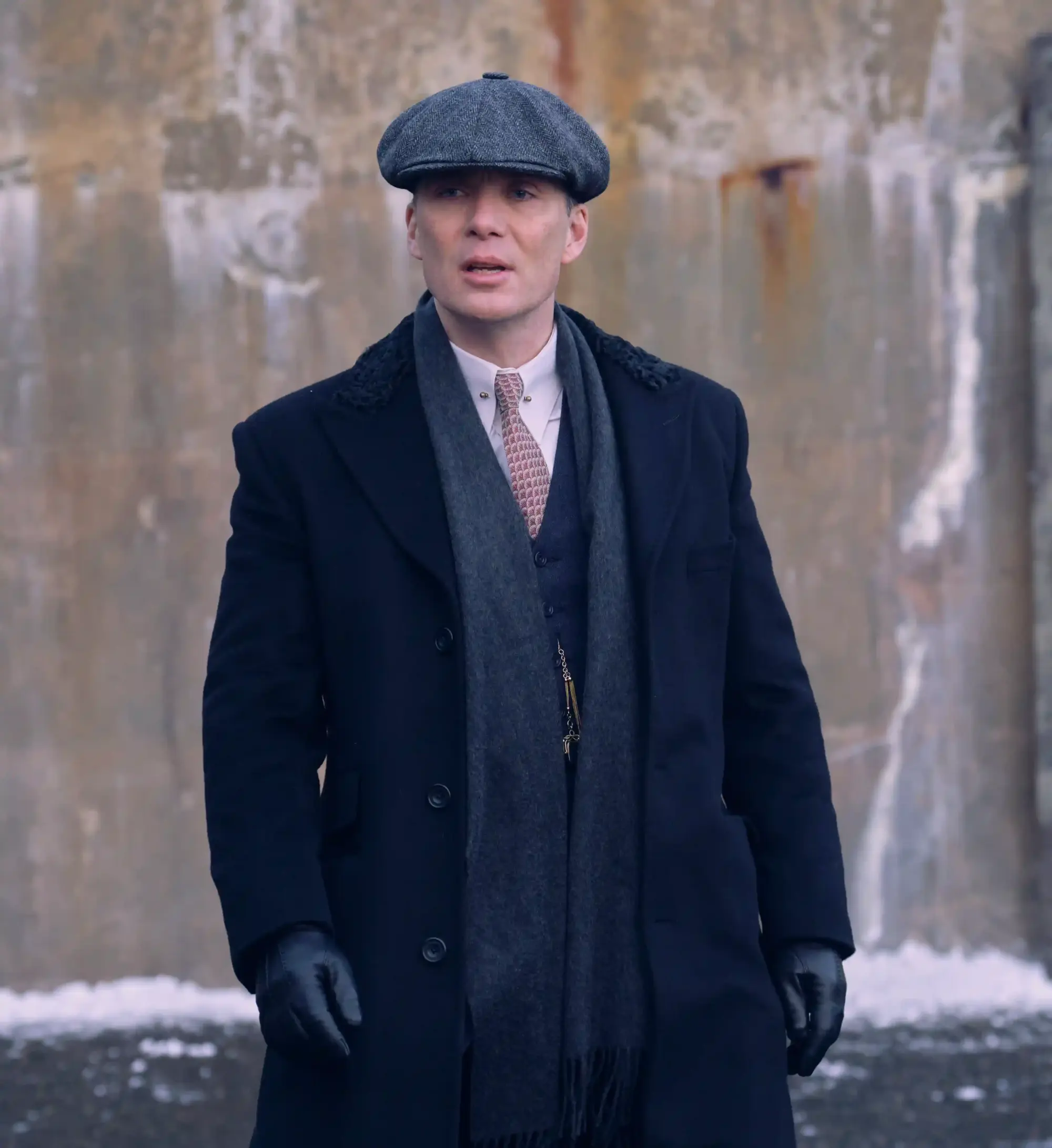 Peaky Blinders'ın başrolü Thomas Shelby, gri tüvit kasketi, yakası kürklü koyu renk paltosu, eşarbı, şık kravatı ve deri eldivenleriyle, 1920'lerin maskülen zarafetini yansıtan güçlü ve etkileyici duruşuyla modern erkek modasında stil sahibi bir gentleman örneği sunuyor.