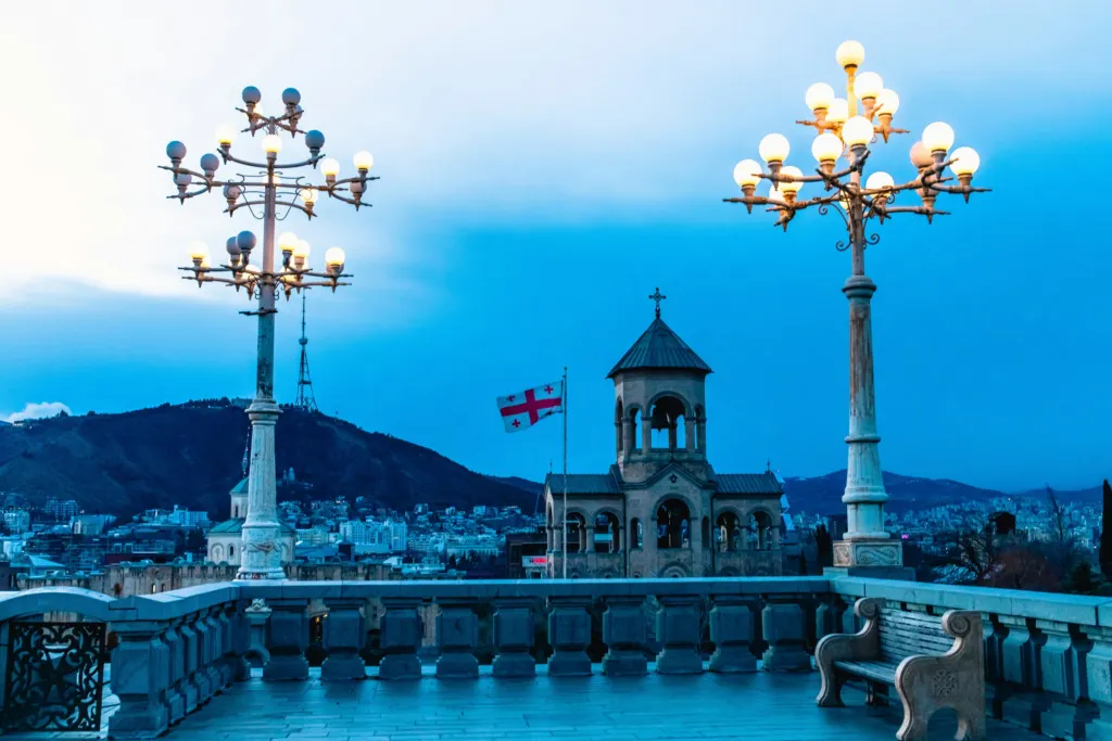 Tiflis'in Sameba Katedrali'nin terasından çekilmiş bu büyüleyici gece manzarasında, Gürcistan bayrağı, kilisenin çan kulesi, şehrin silüeti ve uzaktaki dağlar, ışıklı, çok kollu, işlemeli sokak lambalarının çerçevelediği alacakaranlıkta parlıyor.