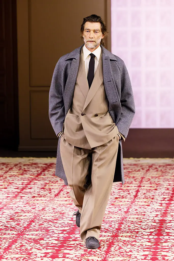 Zegna 2026 sonbahar-kış erkek giyim koleksiyonundan, sakallı bir model, bej tonlarında gevşek kesim kruvaze takım elbisesi ve gri uzun kabanıyla, modern erkek modasında 1920'lerin maskülen zarafetini yansıtan bir görünümle kırmızı desenli bir podyumda yürüyor.