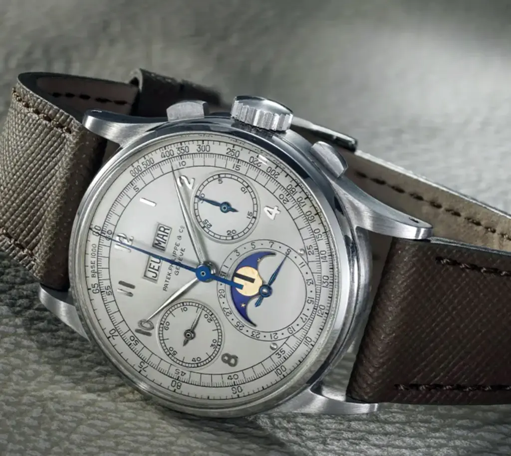 Gri dokulu yüzey üzerinde sergilenen, kahverengi deri kayışlı Patek Philippe vintage kronograf saat; takvim ve ay fazı komplikasyonlarıyla detaylı gümüş kadran tasarımı.