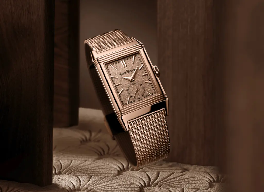 Bronz tonlarda dikdörtgen kasalı, mesh bilezikli lüks Jaeger-LeCoultre Reverso erkek saati dekoratif bir yüzey üzerinde sergileniyor.