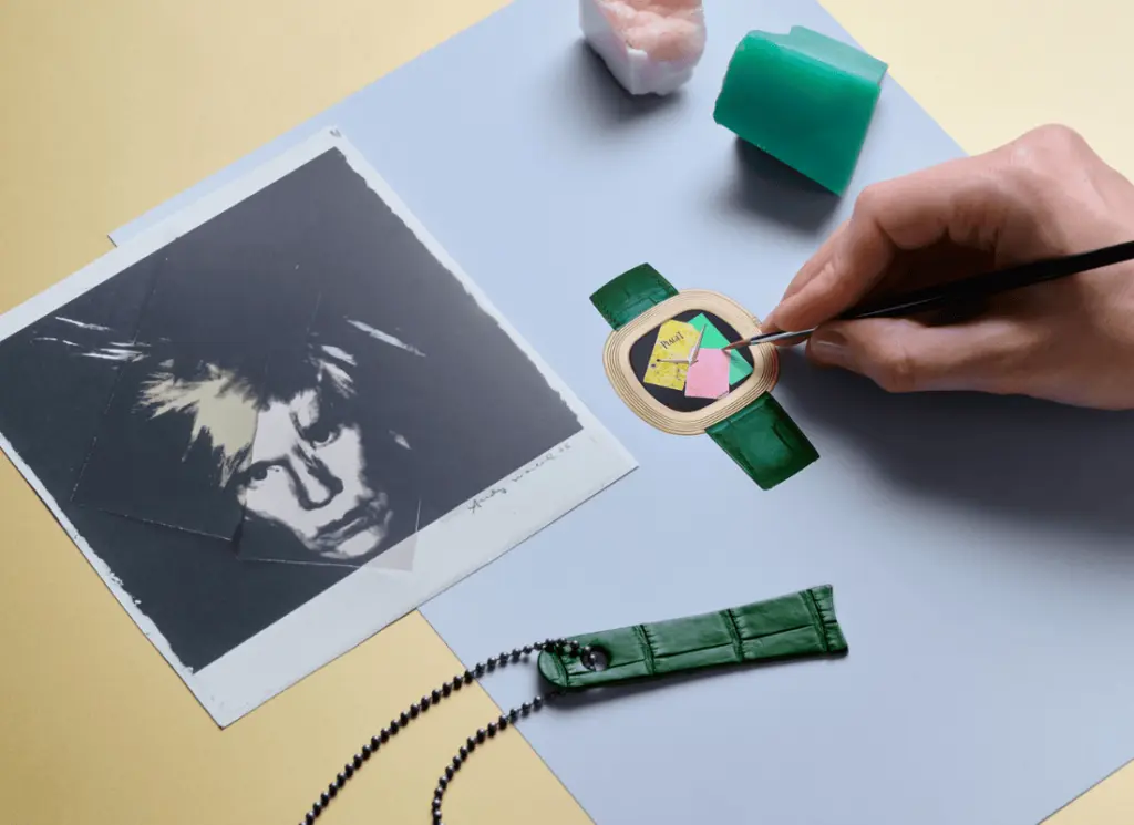 Yeşil kayışlı Piaget saat tasarımının üzerinde çalışan el, fırçayla renkli kolaj detayları eklerken; yanında Andy Warhol portresi ve tasarım malzemeleri bulunan yaratıcı çalışma düzeni.