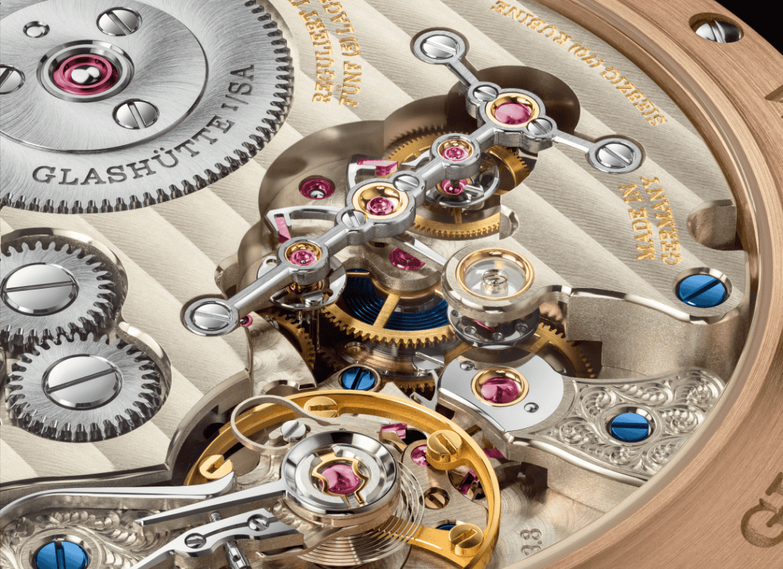 Glashütte kökenli bir mekanik saatin mikroskobik yakın plan görüntüsü; altın, çelik ve mavi çelik vidalarla işlenmiş dişliler, köprüler ve değerli taşlı mekanizma detayları.