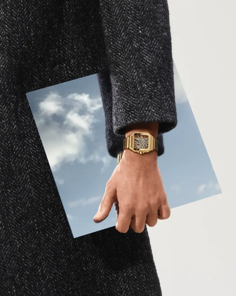 Gold ton metal bilezikli modern tasarım saat takan bir kişinin elinde gökyüzünü yansıtan bir fotoğraf; lüks saat stili ve çağdaş moda detayları.