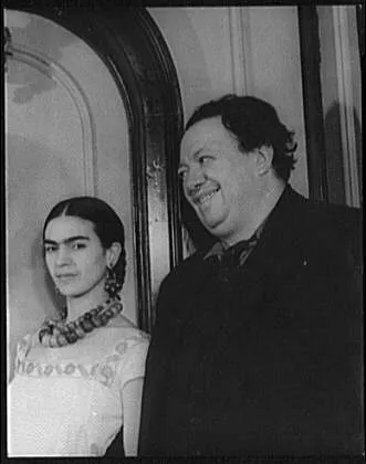 Siyah beyaz fotoğrafta, sol tarafta örgülü saçları, birleşik kaşları ve boncuklu kolyeleriyle Frida Kahlo kameraya bakarken, sağ tarafta neşeli bir ifadeyle gülümseyen Diego Rivera, ikisi de tarihi bir kapı önünde yan yana duruyor.