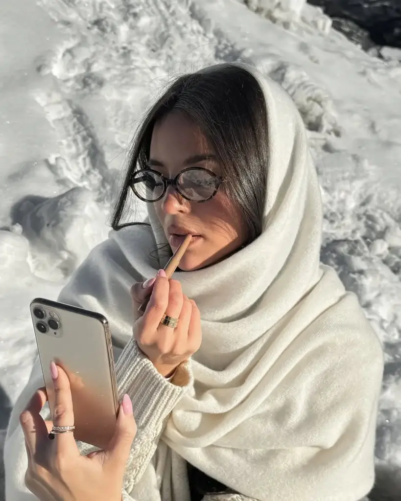 Karlı bir ortamda beyaz şalına sarınmış kadın, telefon ekranına bakarak dudak kalemi ile makyaj yapıyor.