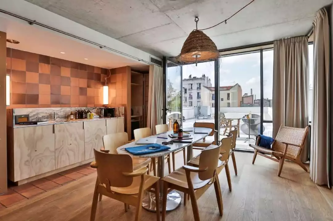 Bu modern yemek odasında, büyük pencerelerden giren doğal ışıkla aydınlanan beyaz oval masa ve etrafındaki açık renk ahşap sandalyeler dikkat çekiyor; tuğla desenli duvar ve hasır avize mekana sıcak bir dokunuş katarken, dışarıdaki şehir manzarası ferah bir atmosfer sunuyor.