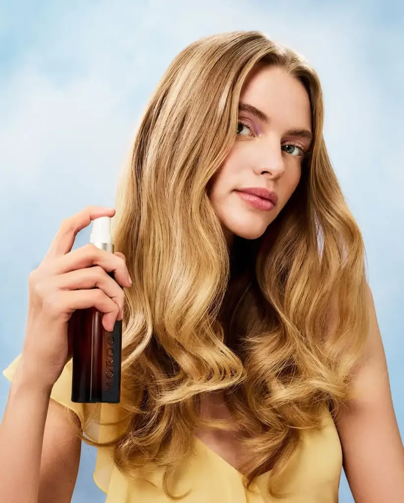 Moroccanoil saç bakım spreyi tutan modelin dalgalı, parlak sarı saçları ve doğal makyajıyla yakın plan çekim.