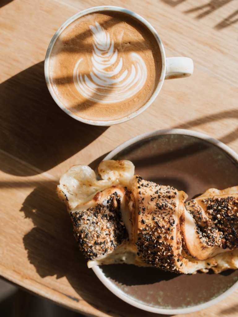 Reykjavik seyahat rehberi kapsamında, güneşli bir ahşap masada, üzerinde zarif latte sanatı bulunan bir fincan kahve ile bolca susam ve haşhaş tohumlarıyla kaplı, çörek benzeri lezzetli bir hamur işi ikilisinin kuşbakışı görünümü, şehrin keyifli kafe kültürünü yansıtıyor.