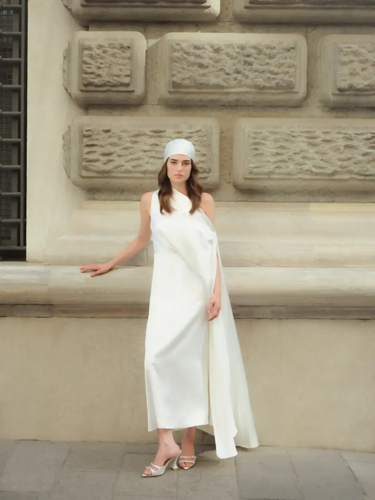 Modern şehir dokusunda poz veren, minimalist beyaz drape elbise giyen model – sade gelinlik trendleri 2026.