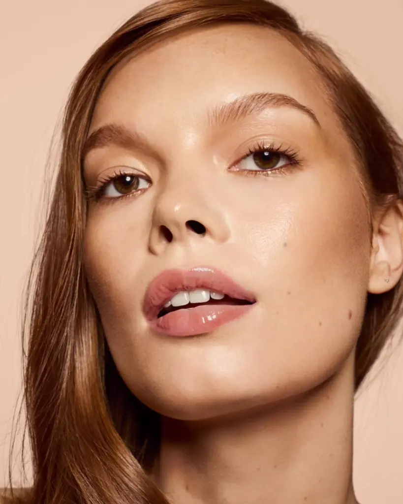 Fenty Beauty ışıltılı ten makyajı ile doğal parlak cilt görünümü, nude gloss dudaklar ve minimal göz makyajına sahip modelin yakın plan yüzü.