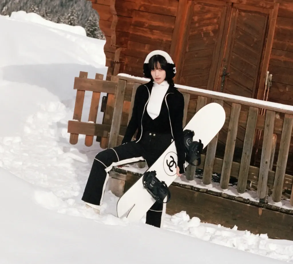 Chanel kayak koleksiyonundan siyah bir tulum giyen ve karlı bir ortamda elinde snowboard ile poz veren model