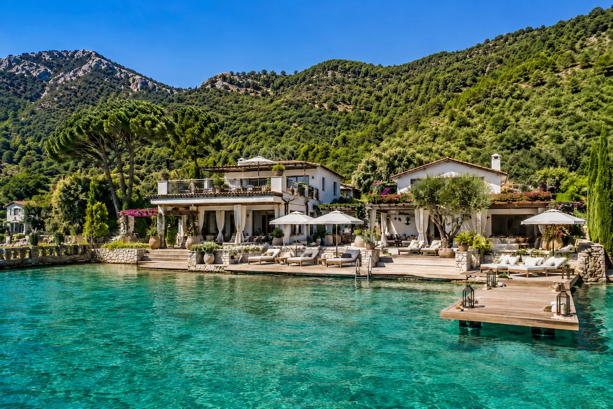 Turkuaz denizin kıyısında konumlanan denize sıfır otel, şezlonglar, iskele ve yemyeşil dağ manzarası